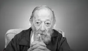 an elderly man sits in an interview during a reflective moment.