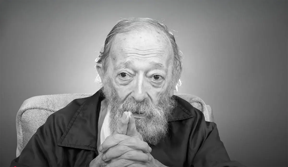 an elderly man sits in an interview during a reflective moment.