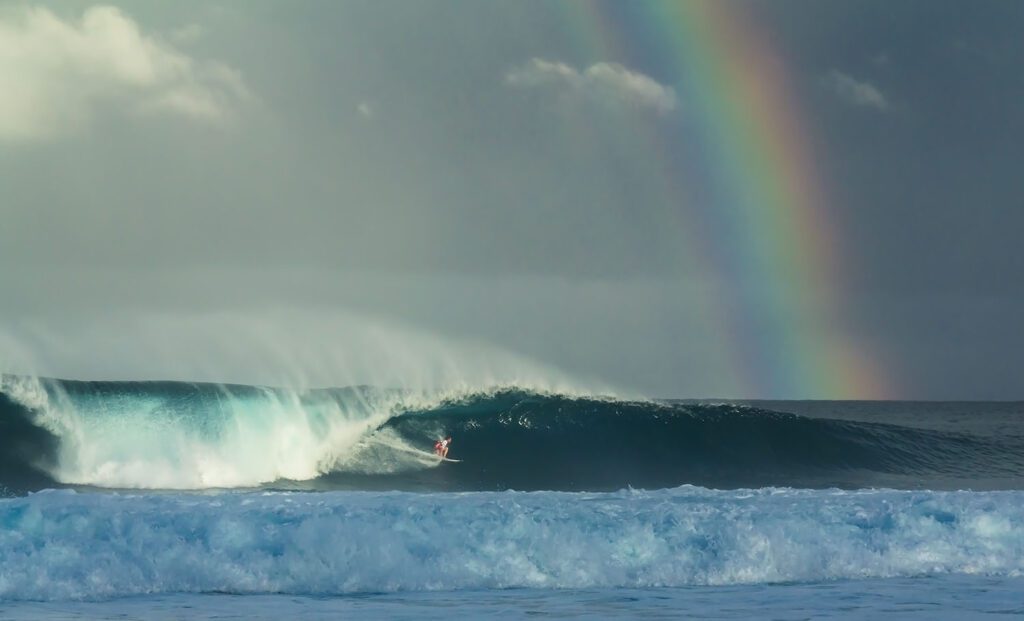 a surfer pulls into the barrel at pipeline with a large rainbow behind it
