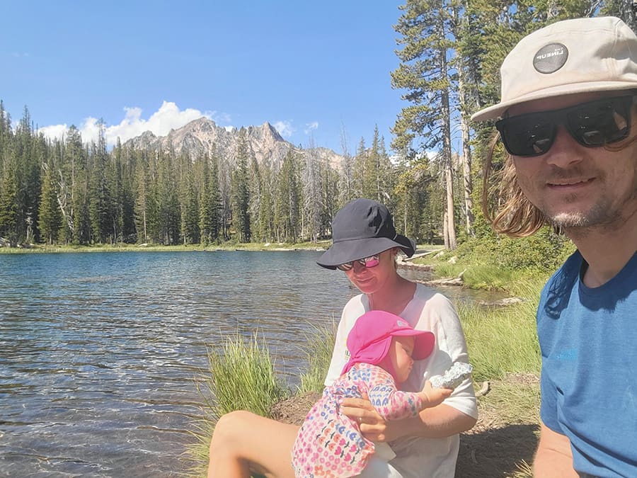 a family of three sit beside an alpine lake in the sawtooth mountains while the baby sleeps.